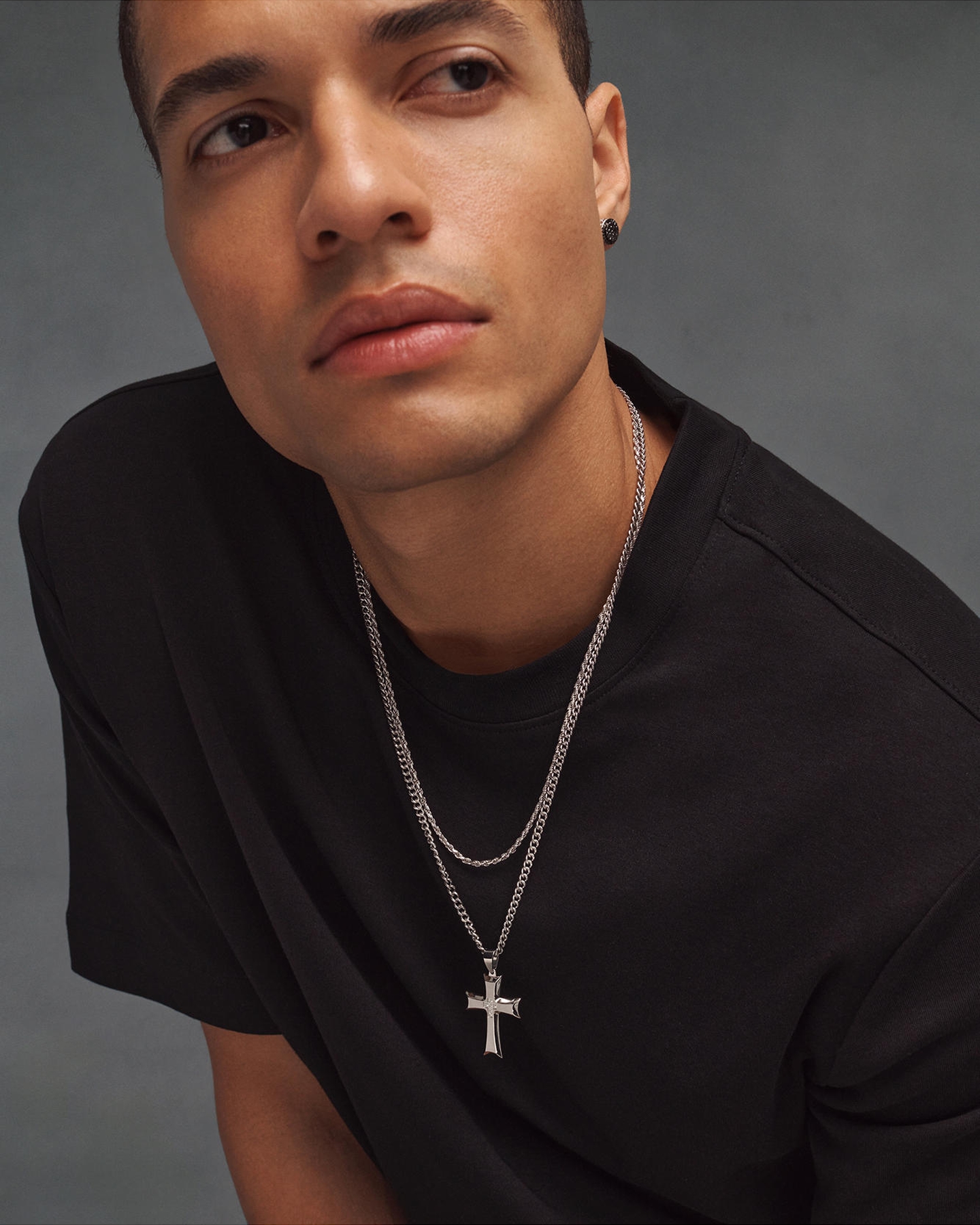 Kay stainless steel cross pendant necklace layered with a polished curb chain, paired with black stud earrings in a modern men's jewelry look