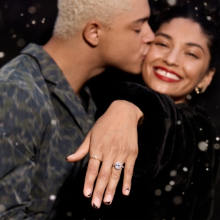 Man kissing woman's cheek while she shows off her engagement ring, smiling