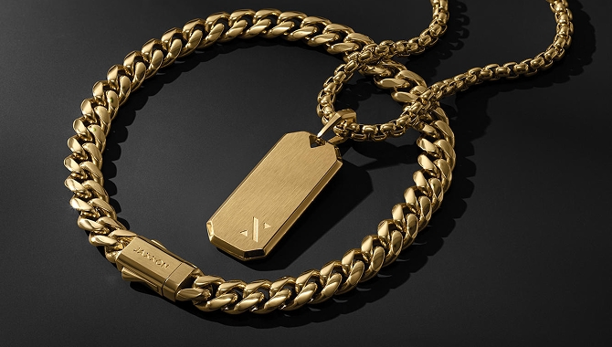 A gold chain bracelet with an attached gold charm, laying on top of another gold chain bracelet.