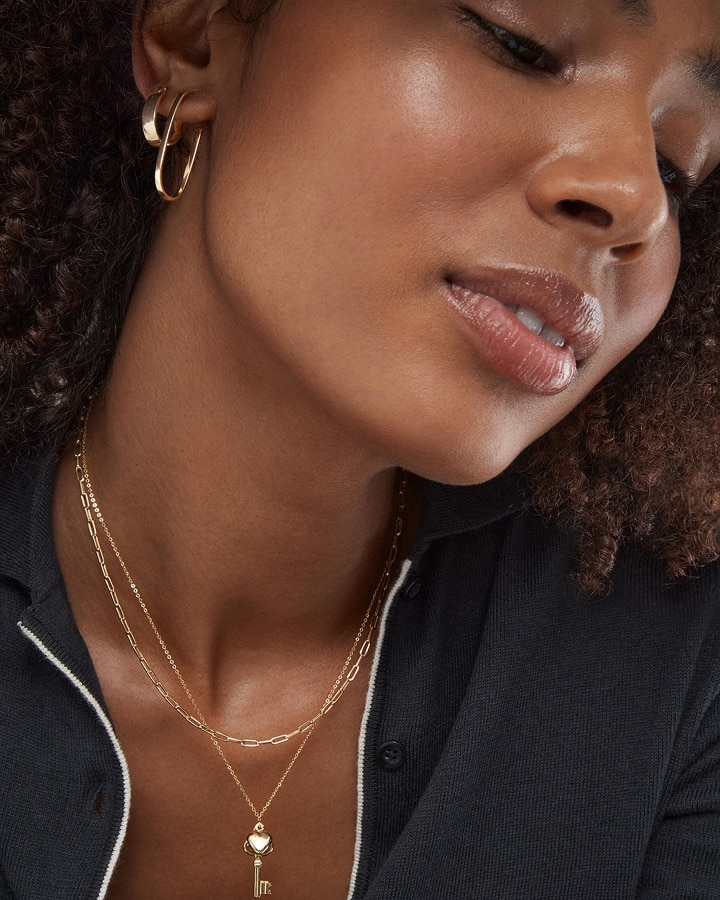 A woman wearing two styles of gold earrings, and two gold chain necklaces from KAY.