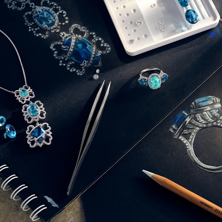 Pencil, tweezers, loose stones, and custom design sketches on black paper next to final product