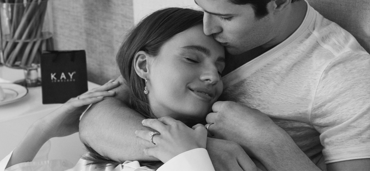 Woman laying on couch with man kissing her forehead with a KAY gift bag next to them