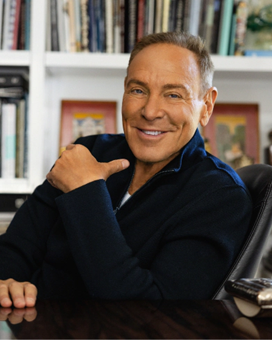 Portrait of jewelry designer Neil Lane seated in a studio surrounded by design books and artwork, representing the inspiration, craftsmanship, and creative vision behind the Kay Neil Lane Artistry jewelry collection.