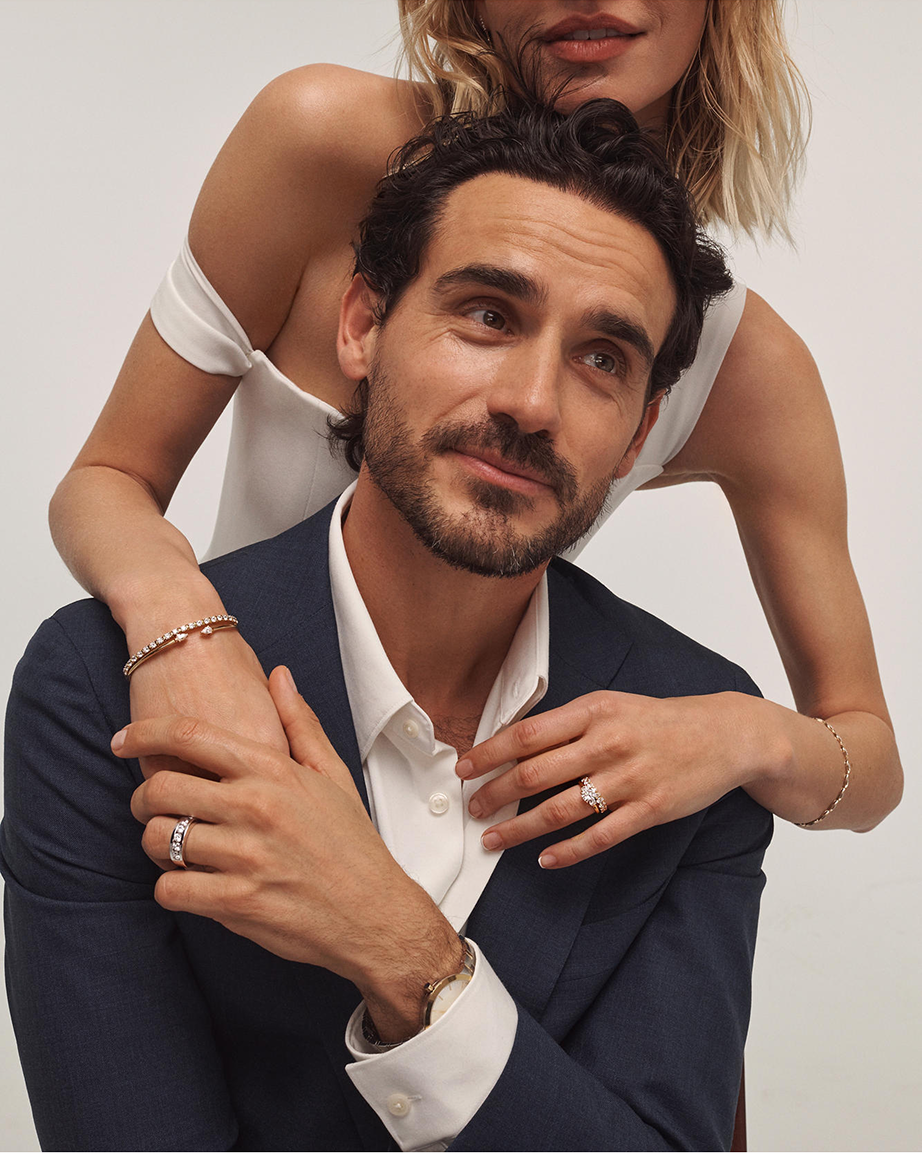 Kay Leo Diamond bridal rings showcased in a luxury lifestyle image, featuring a men's channel-set diamond wedding band and a women's halo diamond engagement ring accented by diamond bracelets and fine jewelry styling
